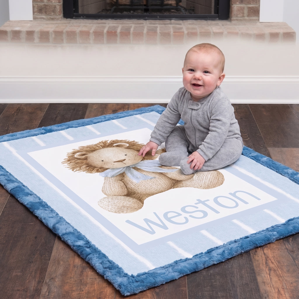 A baby wearing a gray onesie sits on a personalized blue and white blanket featuring a teddy bear and the name 'Weston', placed on a dark wood floor in front of a fireplace.