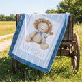 A personalized baby blanket draped over a rustic wooden cart in a grassy outdoor field. The blanket is light blue with a ruffled border and features a central illustration of a beige teddy bear with a blue bow, above the name 'Weston' in light blue text.