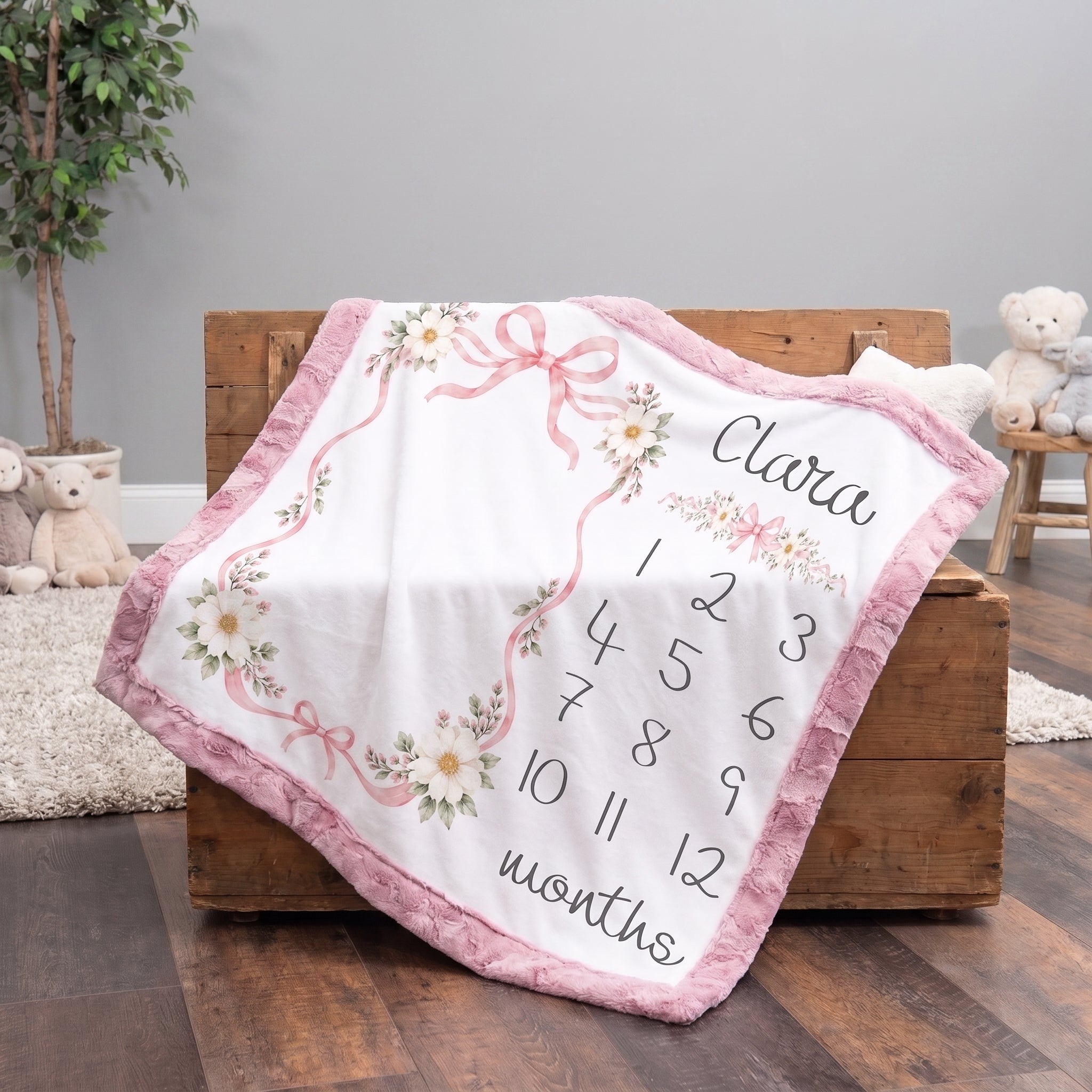 A personalized baby blanket draped over a wooden chest in a nursery setting. The blanket is white with a pink ruffled border, decorated with pink bows, floral patterns, and the name 'Clara' along with numbers 1 through 12 and the word 'months' for customization.