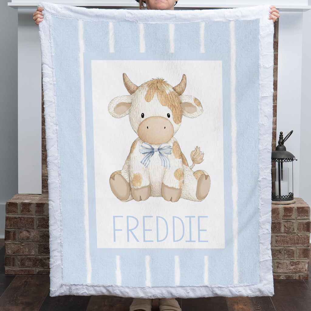 A person holds up a light blue blanket featuring a central illustration of a cow named 'FREDDIE' in blue text. The blanket has white vertical stripes and a white border with fringed edges.