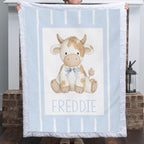 A person holds up a light blue blanket featuring a central illustration of a cow named 'FREDDIE' in blue text. The blanket has white vertical stripes and a white border with fringed edges.