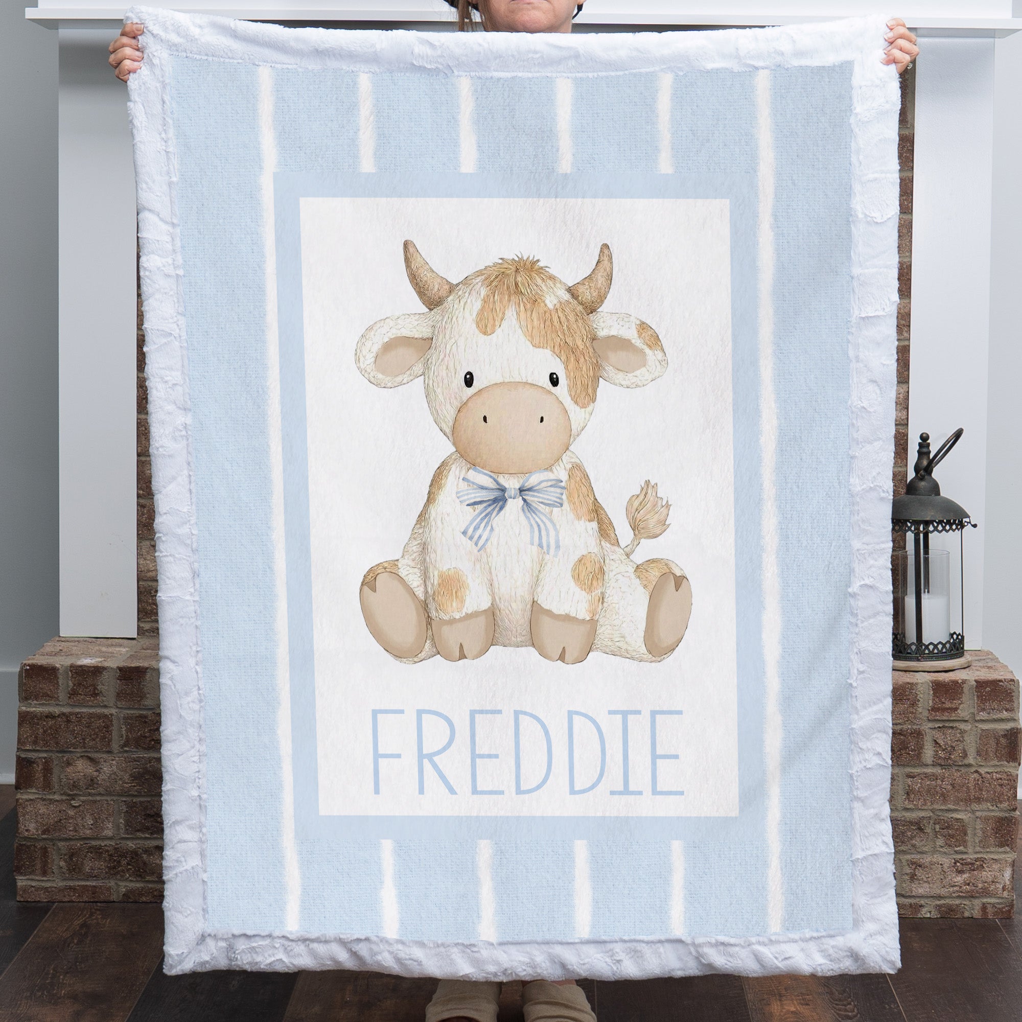 A person holds up a light blue blanket featuring a central illustration of a cow named 'FREDDIE' in blue text. The blanket has white vertical stripes and a white border with fringed edges.