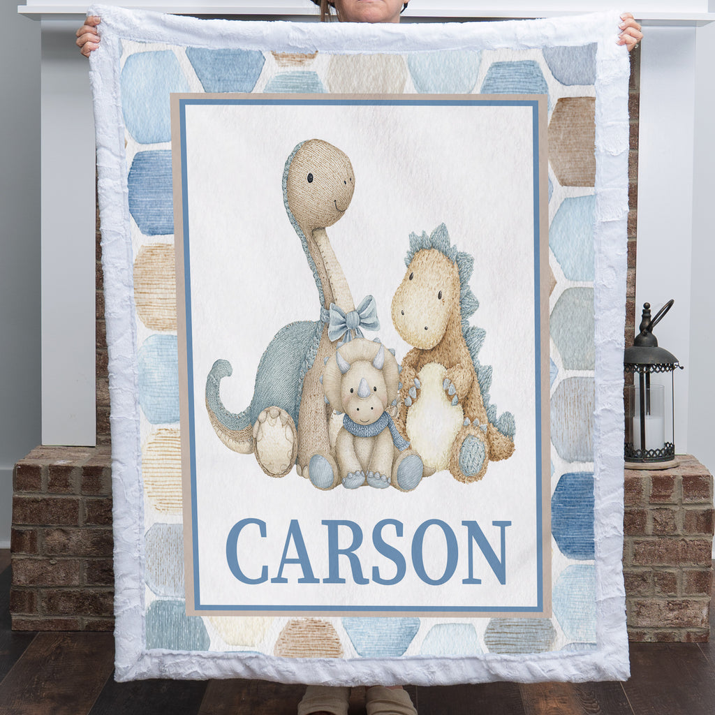 A personalized quilted blanket featuring three embroidered-style dinosaur toys and the name 'CARSON' in blue block letters, held up by a person in front of a fireplace.