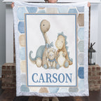 A personalized quilted blanket featuring three embroidered-style dinosaur toys and the name 'CARSON' in blue block letters, held up by a person in front of a fireplace.