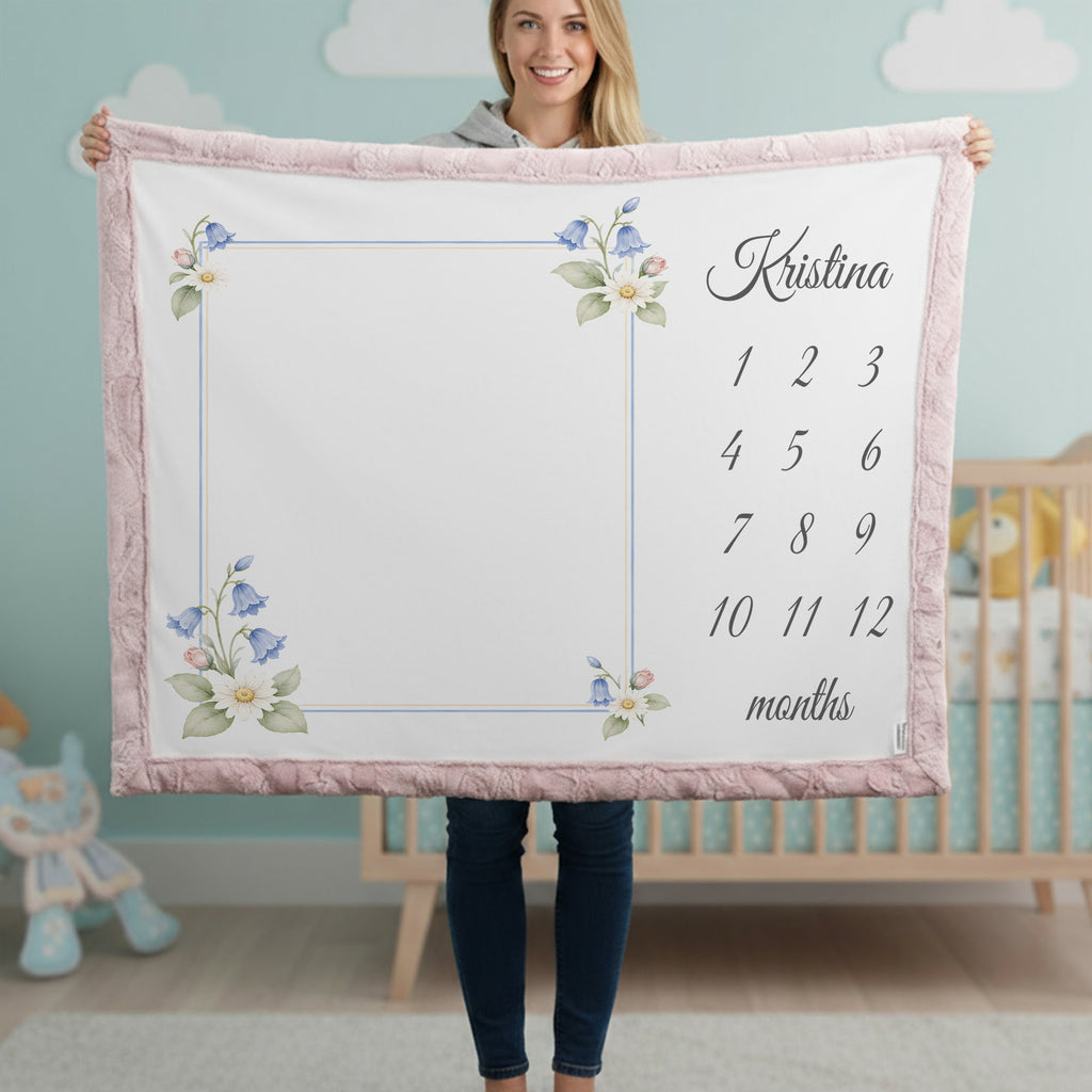 A woman stands in a nursery holding up a personalized baby blanket. The blanket has a white center with a floral border, a blue vertical stripe, and a right side printed with the name 'Kristina' and a 12-month grid.