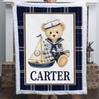 A personalized teddy bear blanket held up by a person in front of a fireplace. The blanket features a navy blue and white plaid border, a central white panel with a cartoon teddy bear in a sailor outfit holding a sailboat, and the name 'CARTER' printed in navy blue text at the bottom.