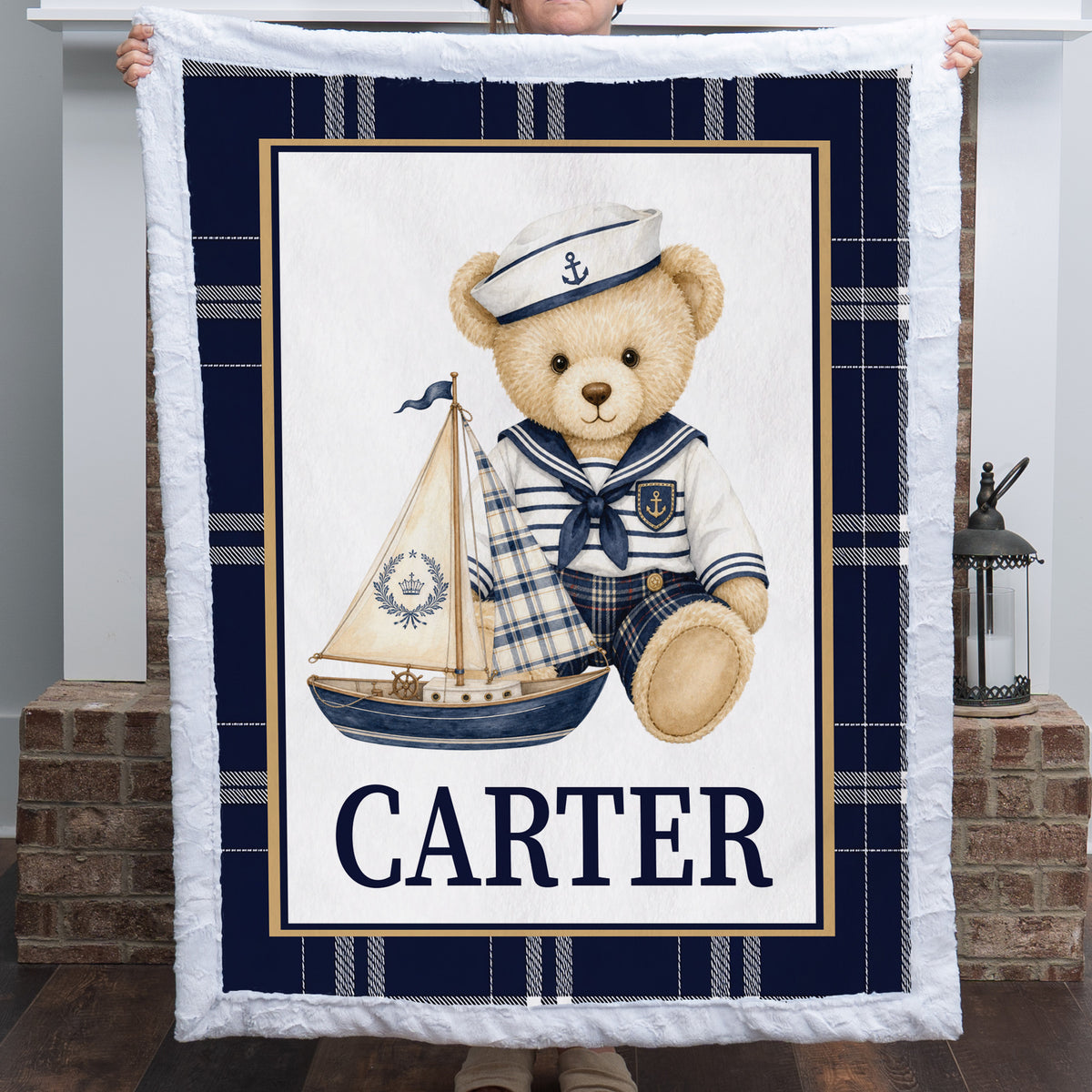 A personalized teddy bear blanket held up by a person in front of a fireplace. The blanket features a navy blue and white plaid border, a central white panel with a cartoon teddy bear in a sailor outfit holding a sailboat, and the name 'CARTER' printed in navy blue text at the bottom.