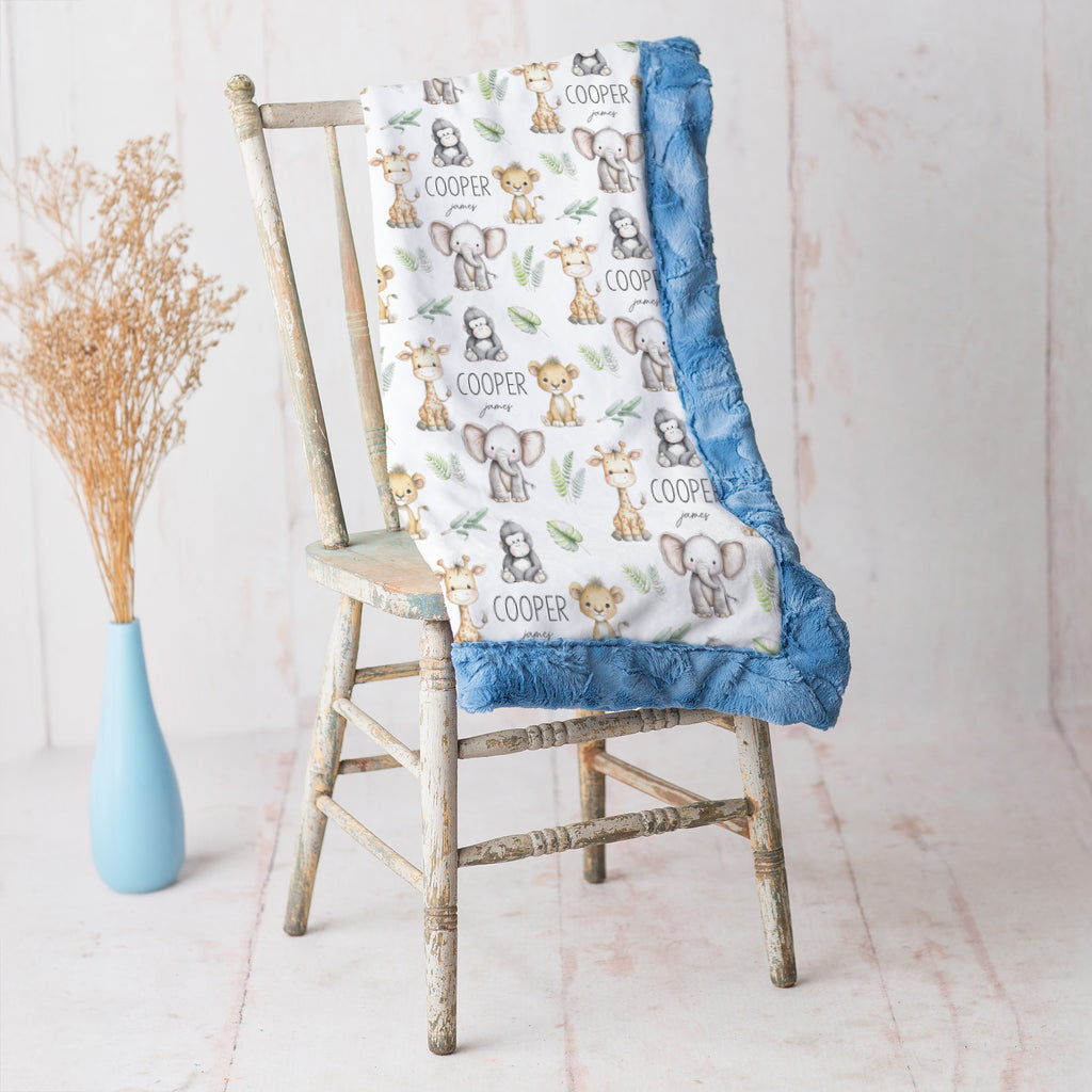 A baby blanket with a white background printed with cartoon safari animals and the name 'COOPER', finished with a blue ruffled trim, draped over a rustic wooden chair next to a light blue vase with dried grass.