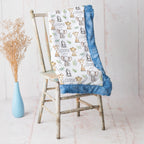 A baby blanket with a white background printed with cartoon safari animals and the name 'COOPER', finished with a blue ruffled trim, draped over a rustic wooden chair next to a light blue vase with dried grass.