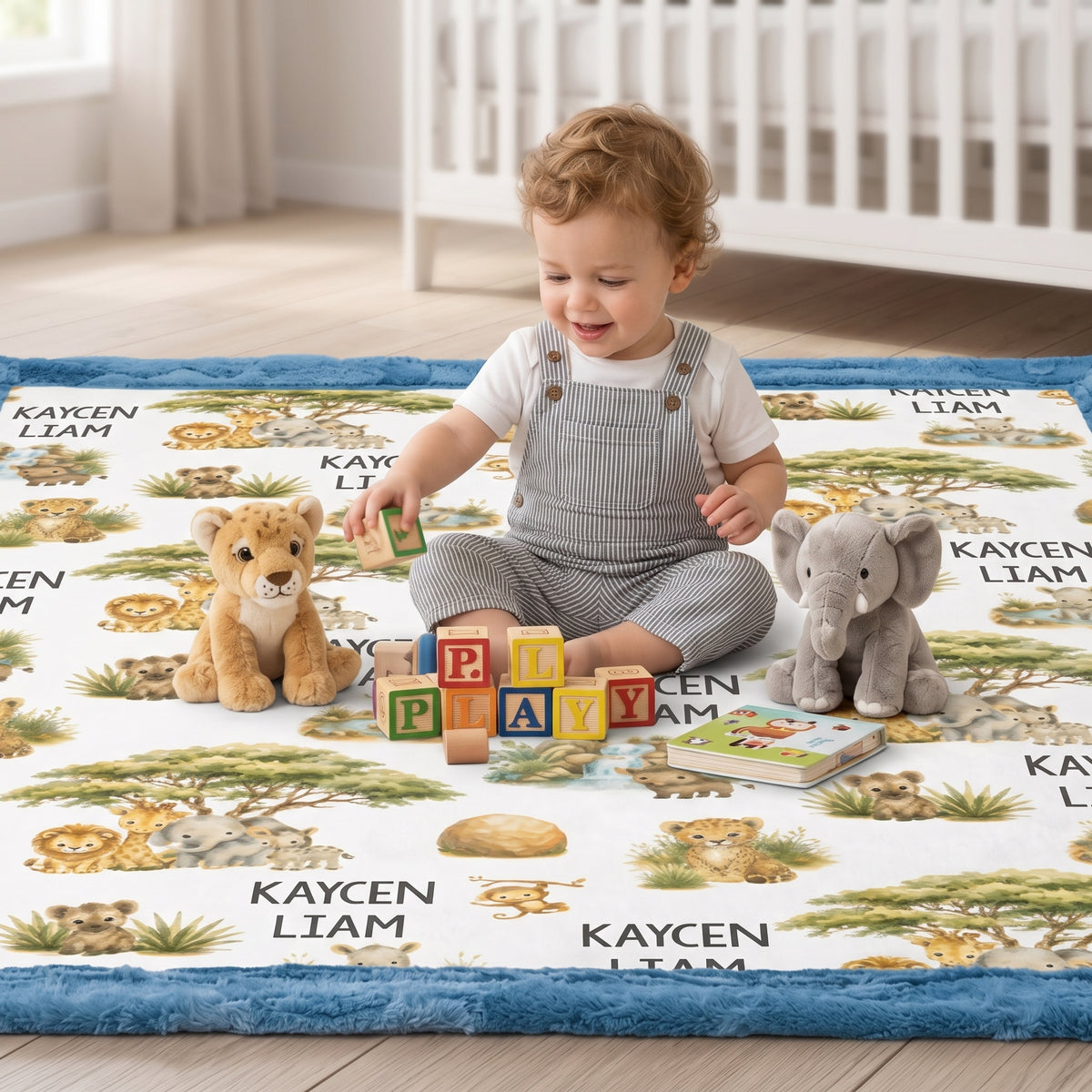 A toddler sits cross-legged on a safari-themed baby play mat in a bright nursery, surrounded by plush toys and colorful building blocks. The play mat features illustrated animals and the name 'KAYSEN LIAM'.