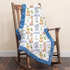 Personalized baby blanket with a white background, blue minky fur trim, and a print of cartoon giraffes, lions, and elephants with the name 'HAYDEN John'. The blanket is draped over a rustic wooden rocking chair on a dark wood floor against a beige wall.