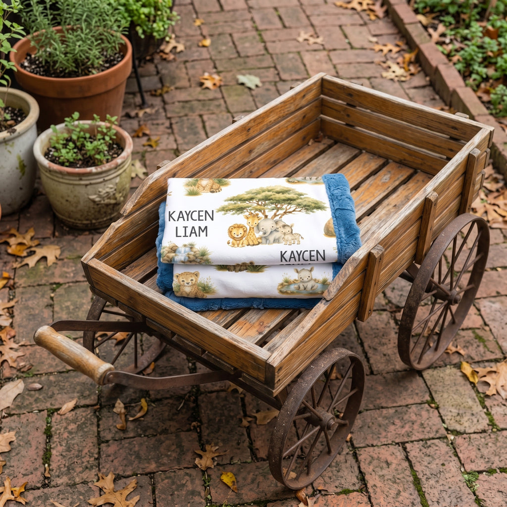 A personalized baby blanket with a safari lion print and the names 'KAYCEN LIAM' is folded and displayed inside a rustic wooden toy cart on a brick patio.