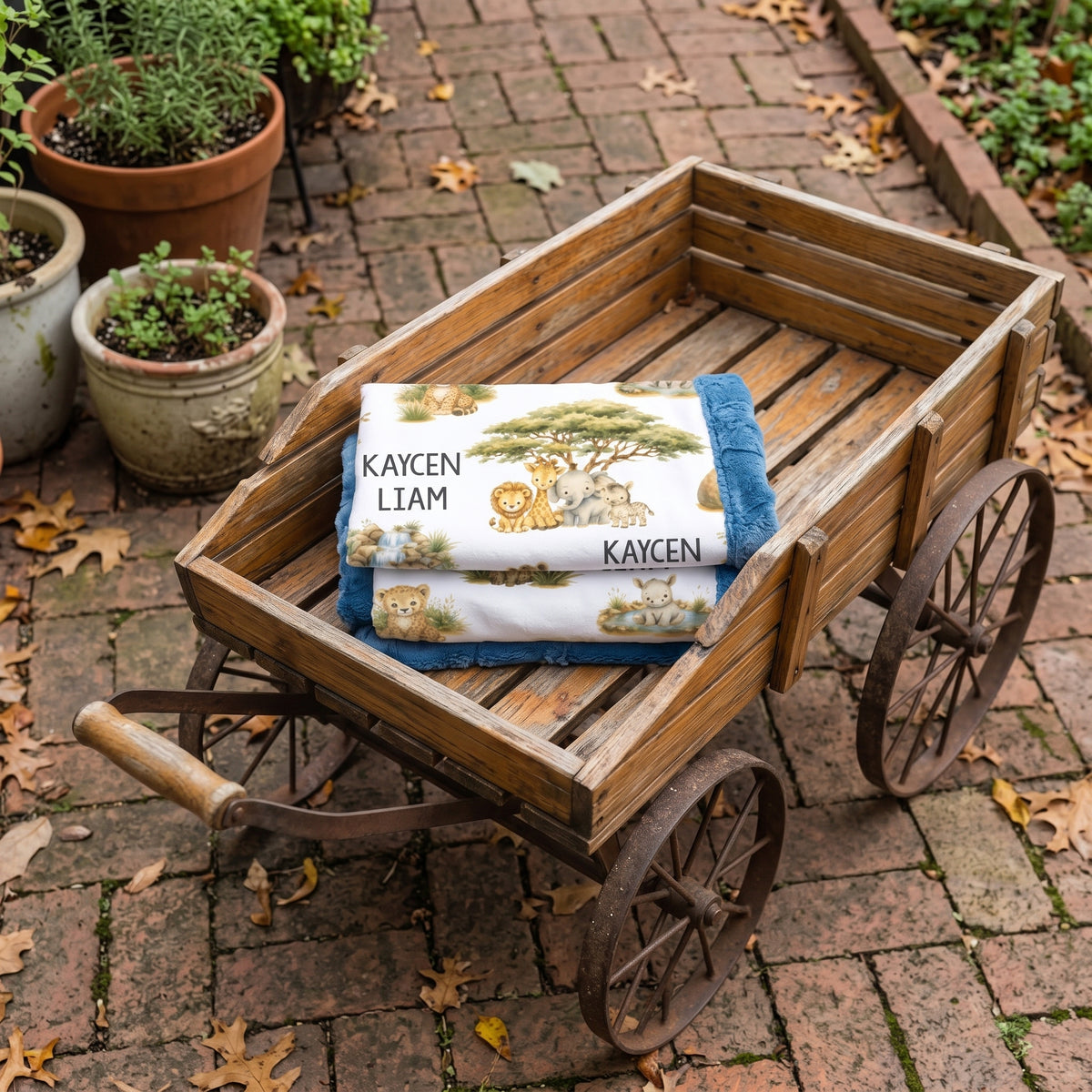 A personalized baby blanket with a safari lion print and the names 'KAYCEN LIAM' is folded and displayed inside a rustic wooden toy cart on a brick patio.