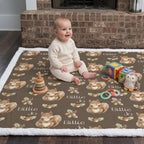 A baby sits on a brown play mat with a white sherpa border, featuring a repeating pattern of brown squirrels, acorns, and the text 'Billie Jo'. The baby is wearing a cream knit outfit and is surrounded by toys in a living room setting in front of a brick fireplace.