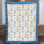 A personalized baby blanket held up by a child, featuring a white center printed with giraffes, elephants, lions, and the name 'HAYDEN John' in blue, surrounded by a blue ruffled border.
