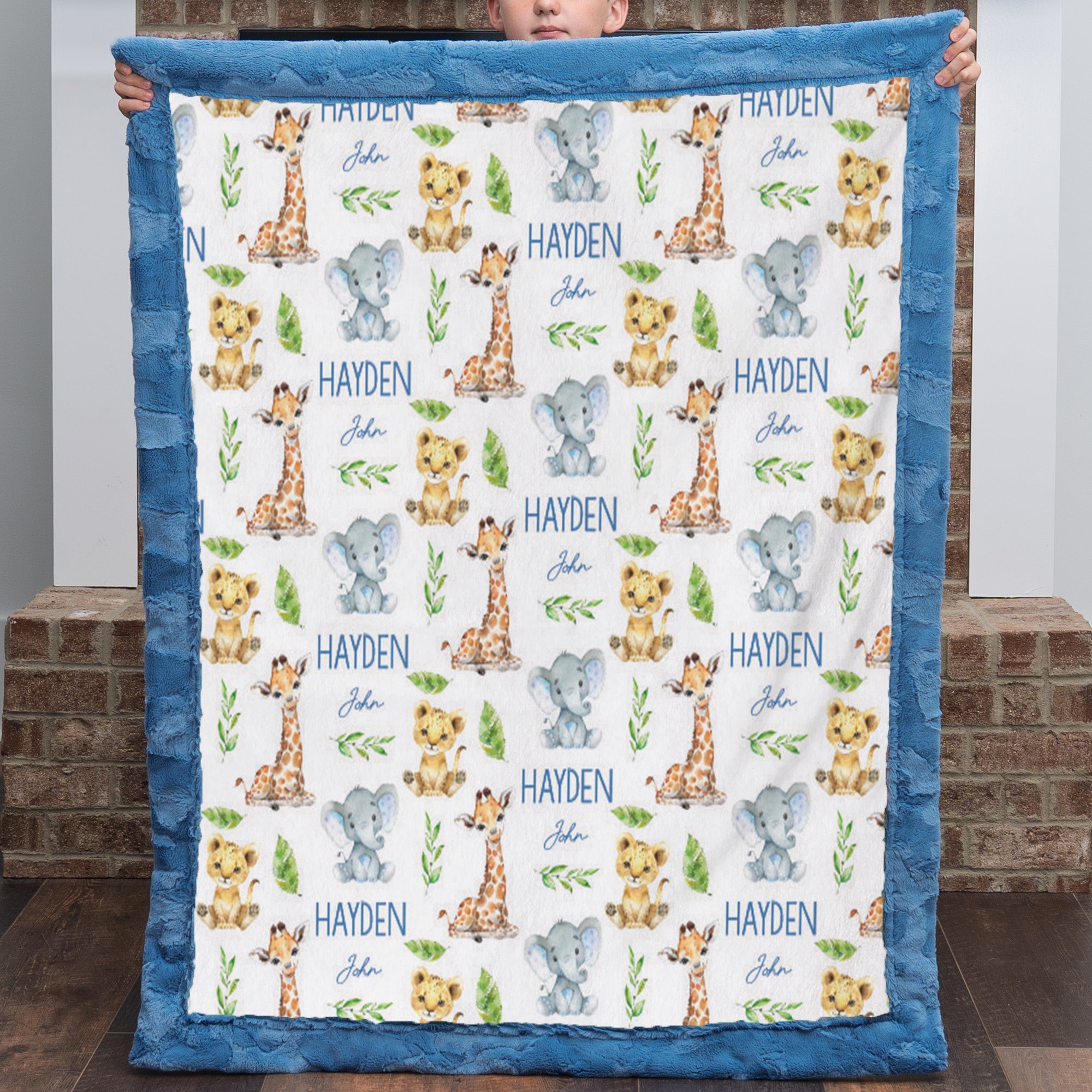 A personalized baby blanket held up by a child, featuring a white center printed with giraffes, elephants, lions, and the name 'HAYDEN John' in blue, surrounded by a blue ruffled border.