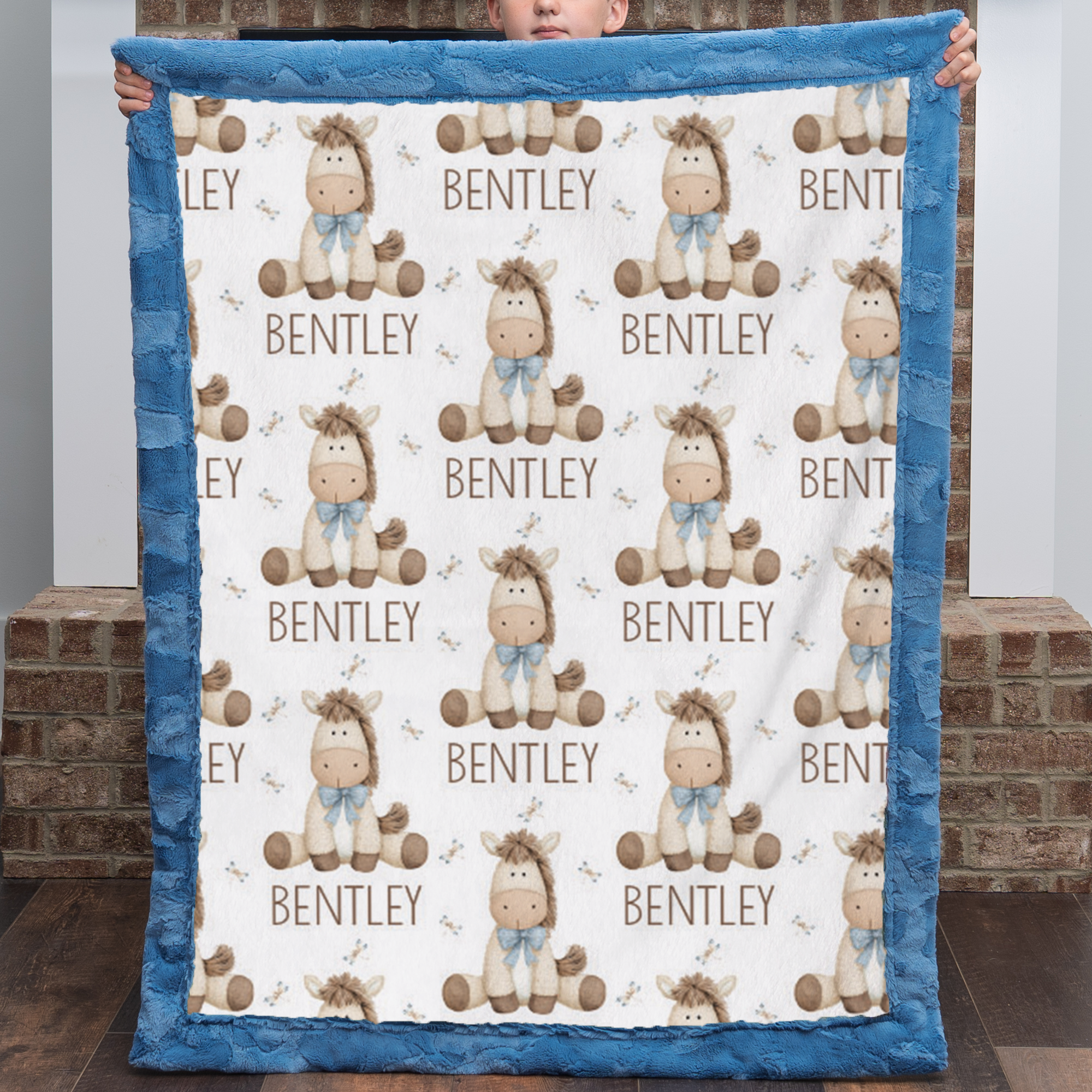 A personalized baby blanket with a blue minky fabric border and a white center printed with cartoon horses, the name 'BENTLEY', and small antler motifs. A child is holding the blanket up in front of a brick wall.