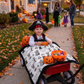 A young child wearing a pirate hat sits in a red wagon on a Halloween-themed front yard path, with pumpkins, candy baskets, and other children in costumes in the background.