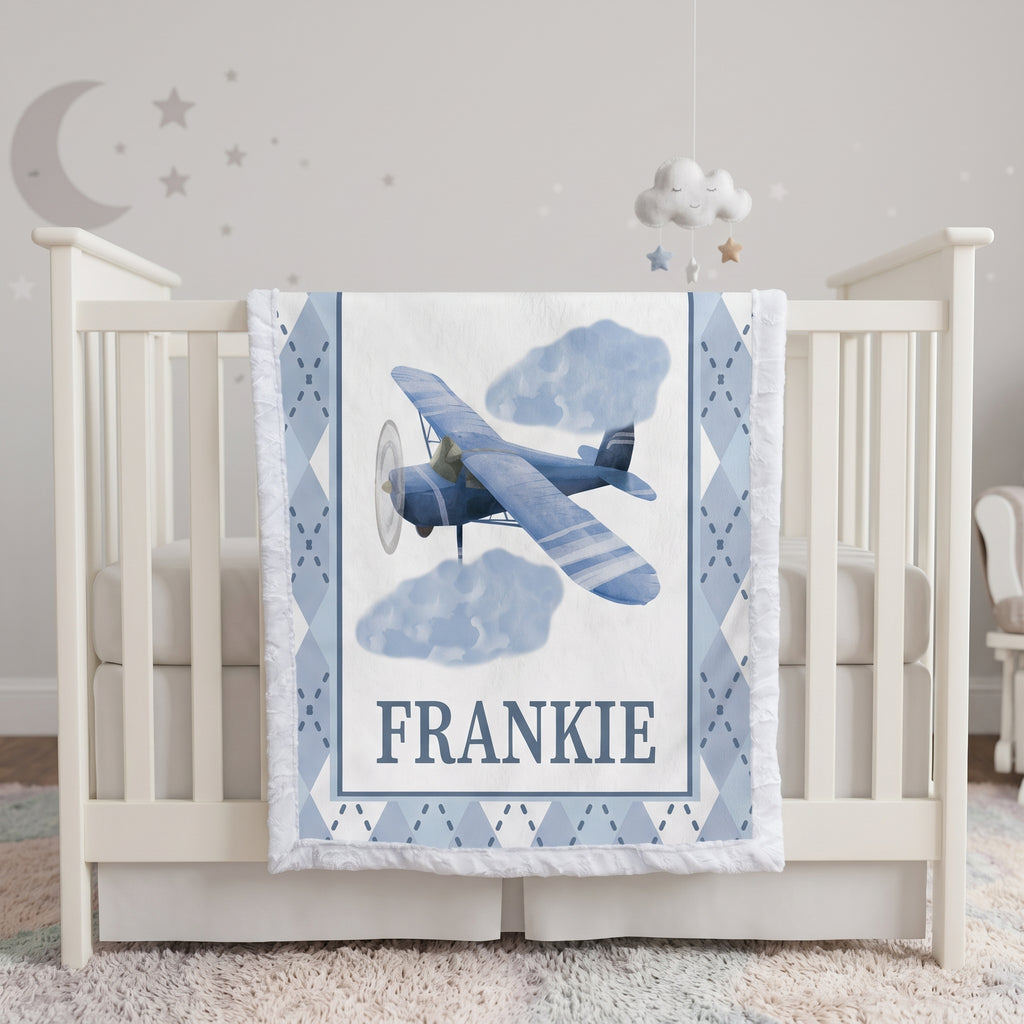 A personalized baby crib bedding set featuring a blue vintage biplane and clouds on a white background with the name 'FRANKIE' printed in navy. The crib is white with a matching blue and white argyle quilted bumper, set in a softly lit nursery with a cloud mobile and wall decals.