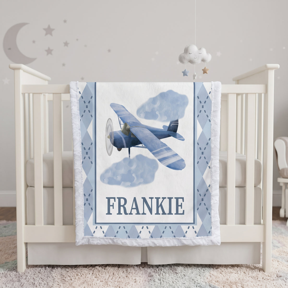 A personalized baby crib bedding set featuring a blue vintage biplane and clouds on a white background with the name 'FRANKIE' printed in navy. The crib is white with a matching blue and white argyle quilted bumper, set in a softly lit nursery with a cloud mobile and wall decals.