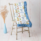 Personalized baby blanket with a safari animal print and blue ruffled trim, draped over a rustic wooden chair next to a light blue vase with dried grass.