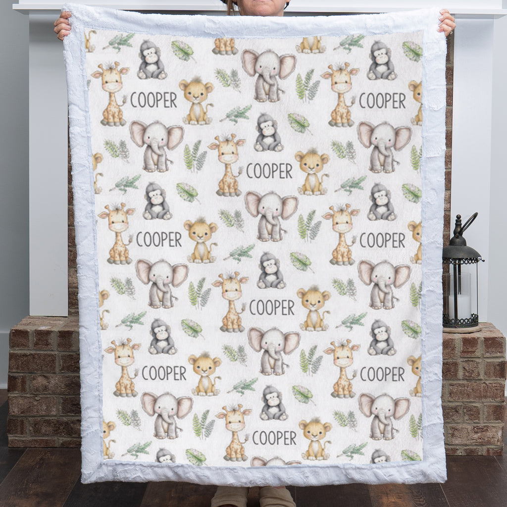 A person holds up a square quilted baby blanket featuring a safari animal print with giraffes, elephants, monkeys, and the name 'COOPER' repeated on an off-white background.