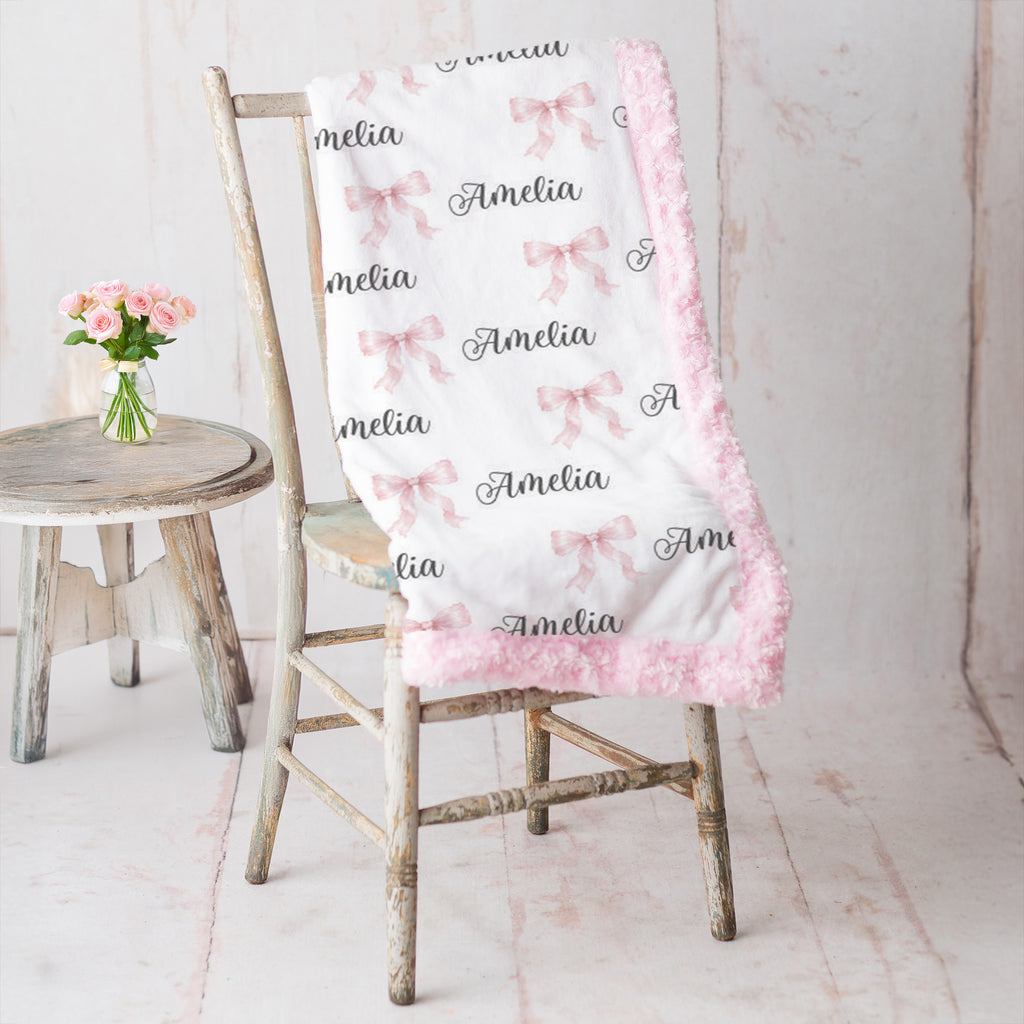 A white baby blanket with pink fuzzy trim, printed with pink bows and the name 'Amelia' in black script, draped over a rustic wooden chair next to a small stool with pink flowers.