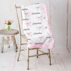 A white baby blanket with pink fuzzy trim, printed with pink bows and the name 'Amelia' in black script, draped over a rustic wooden chair next to a small stool with pink flowers.