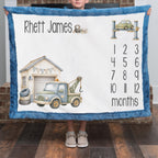 A child holds up a personalized minky blanket for a baby boy. The blanket features a blue fuzzy border, a white center, the name 'Rhett James', a tow truck illustration, a garage, and a 12-months countdown calendar.