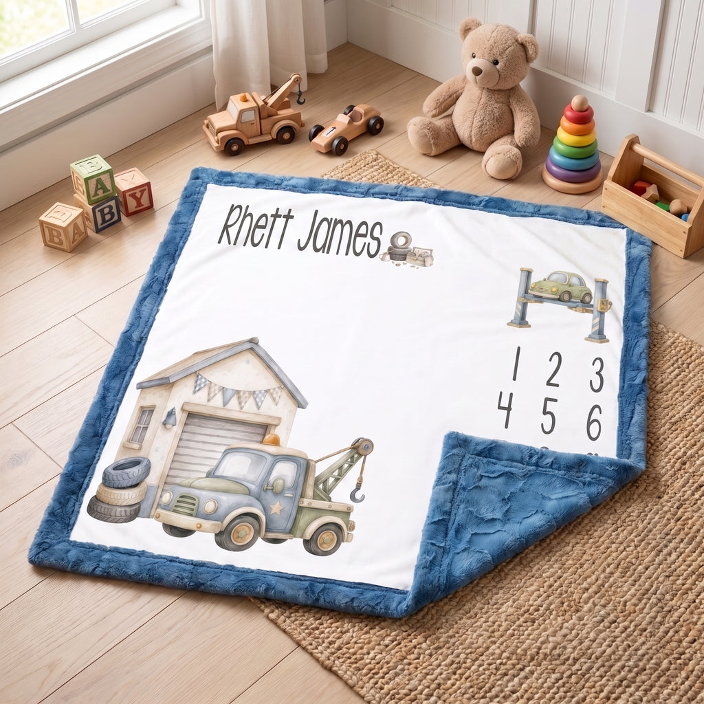 A personalized baby blanket with a blue plush border, featuring a white center printed with a garage truck illustration, the name 'Rhett James', and numbers 1 through 6. The blanket is styled on a woven rug on a wooden floor with toys and a teddy bear in the background.