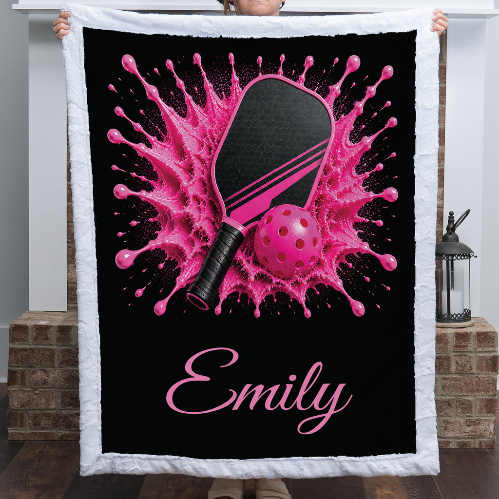 A white fleece blanket with a black center panel featuring a pink and black paddle and ball graphic, with the name 'Emily' in pink script, held up by a person in a home setting.