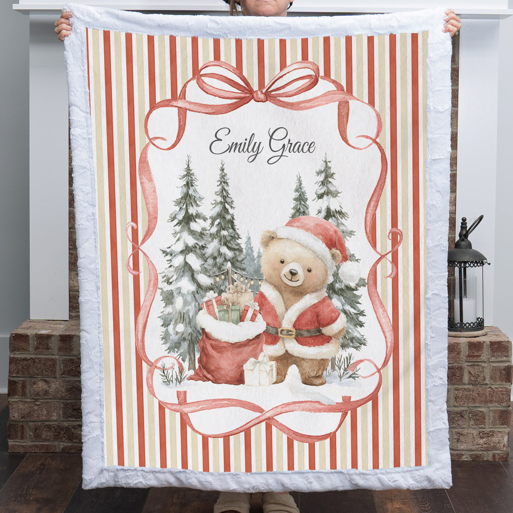 A person holds up a personalized Christmas fleece blanket featuring a teddy bear in a Santa hat, a red gift sack, snow-covered pine trees, and the name 'Emily Grace' in script. The blanket has a white quilted border and red-and-cream vertical stripes, shown in front of a brick fireplace.
