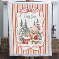 A person holds up a personalized Christmas fleece blanket featuring a teddy bear in a Santa hat, a red gift sack, snow-covered pine trees, and the name 'Emily Grace' in script. The blanket has a white quilted border and red-and-cream vertical stripes, shown in front of a brick fireplace.