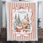 A person holds up a personalized Christmas fleece blanket featuring a teddy bear in a Santa hat, a red gift sack, snow-covered pine trees, and the name 'Emily Grace' in script. The blanket has a white quilted border and red-and-cream vertical stripes, shown in front of a brick fireplace.