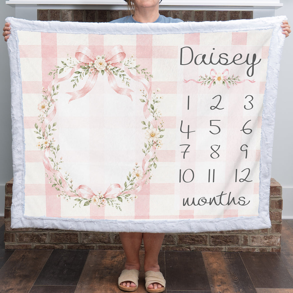A person holds up a personalized baby blanket featuring a pink and white gingham check pattern, a floral wreath design, and a customizable name 'Daisy' with numbered months from 1 to 12. The blanket has a soft white plush border.
