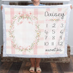 A person holds up a personalized baby blanket featuring a pink and white gingham check pattern, a floral wreath design, and a customizable name 'Daisy' with numbered months from 1 to 12. The blanket has a soft white plush border.
