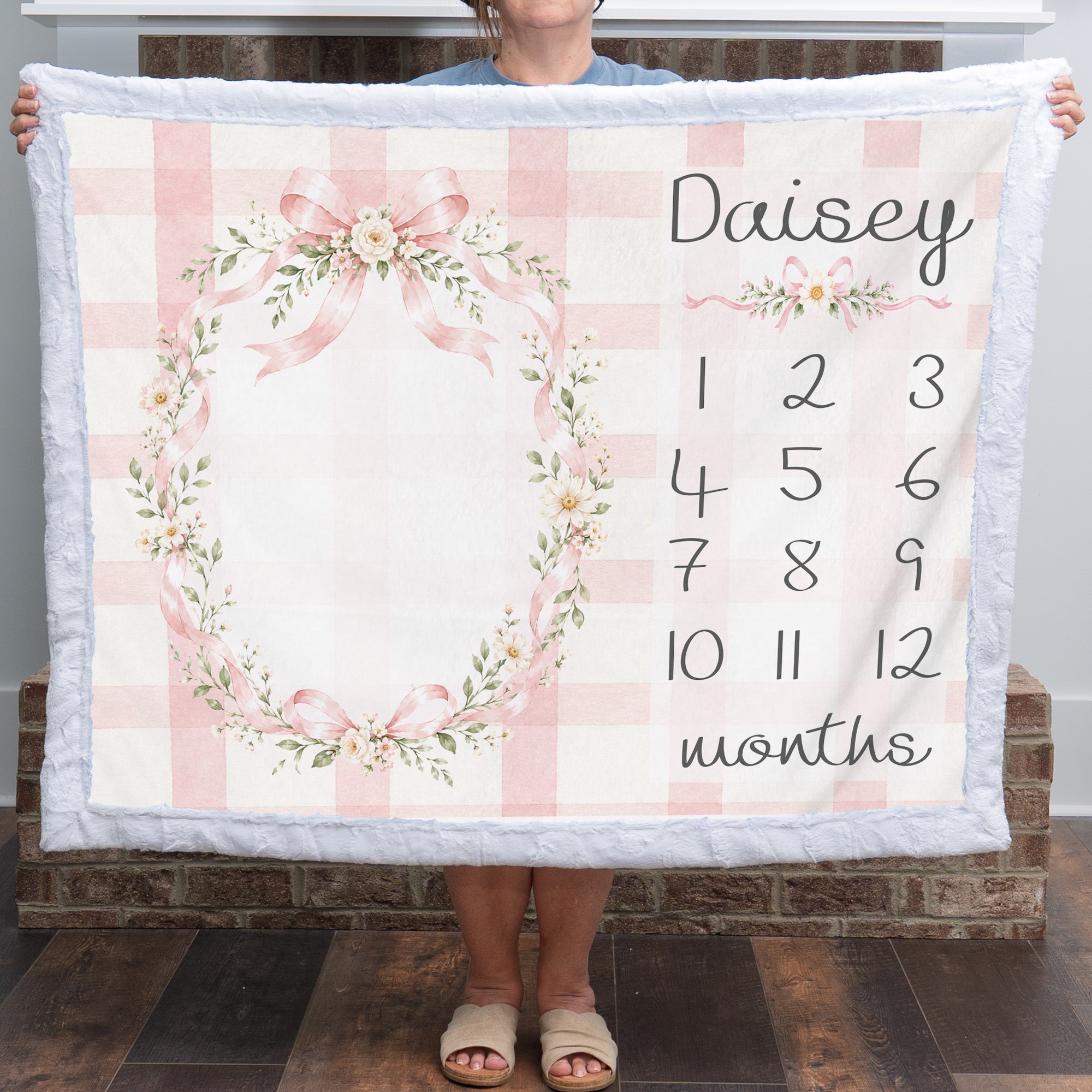 A person holds up a personalized baby blanket featuring a pink and white gingham check pattern, a floral wreath design, and a customizable name 'Daisy' with numbered months from 1 to 12. The blanket has a soft white plush border.